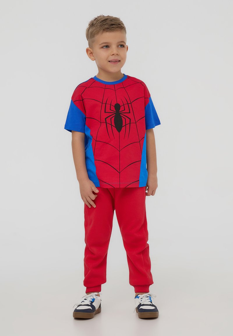 T-shirt a tema Spider-Man rosso e blu con grafica dello spider nera, abbinata a pantaloni rossi. Il tessuto sembra cotone con una texture liscia.