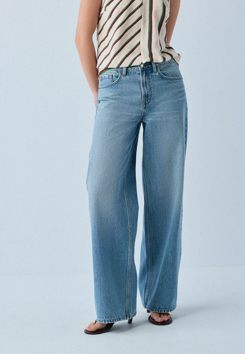 Personne portant un jean large bleu clair, un top rayé sans manches et des sandales à talons noires, debout devant un fond clair uni.