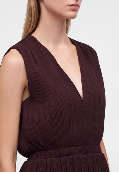 Femme portant une robe plissée sans manches violet foncé avec un profond décolleté en V et une taille cintrée, présentée de l'épaule au milieu du torse sur un fond clair.