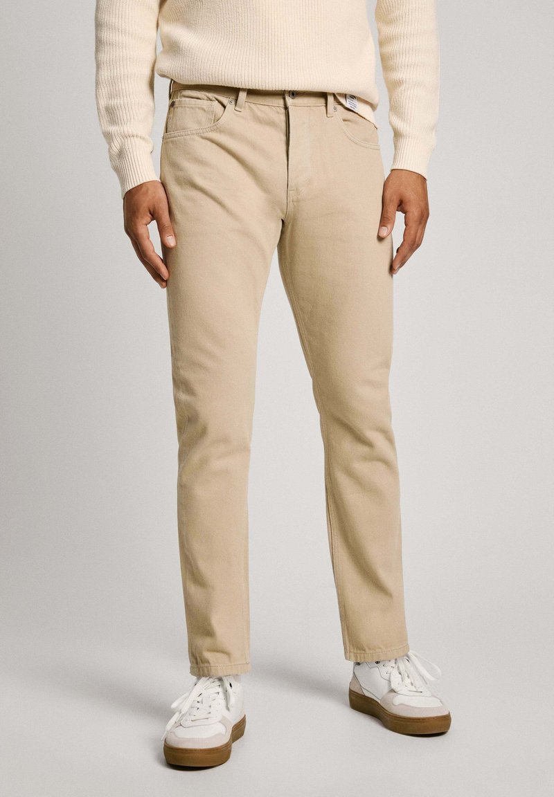 Pantalones beige de algodón con corte recto, diseño de cinco bolsillos y detalles de costura sutiles. Combinados con zapatillas blancas.