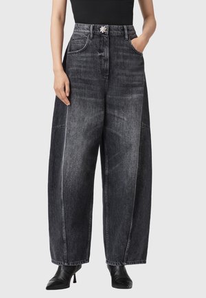 Person trägt hoch taillierte, weit geschnittene schwarze ausgewaschene Jeans mit dekorativem silbernem Knopf, ein schwarzes Oberteil und schwarze Stiefel mit Absatz, steht mit Händen in den Taschen.