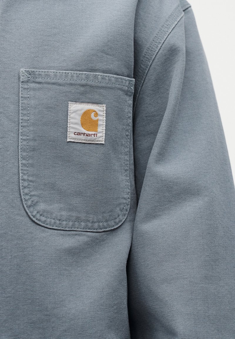Primo piano di una tasca in tessuto Carhartt blu con il logo del marchio visibile cucito nella parte superiore centrale.