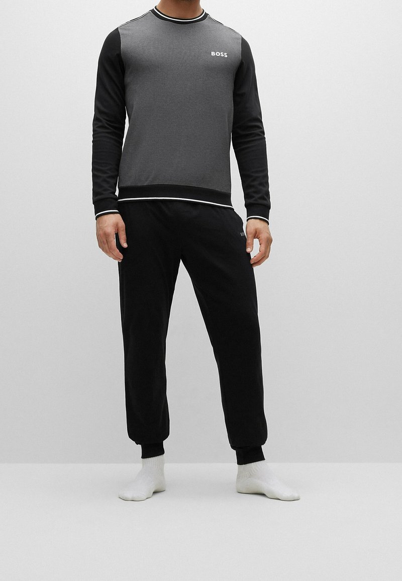 Homme portant un sweat-shirt BOSS à manches longues noir et gris et un pantalon de jogging noir, debout pieds nus sur un fond gris uni.