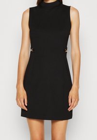 Robe noire sans manches avec un col montant. Elle présente des accents en métal doré à la taille et une silhouette ajustée. Texture du tissu lisse.