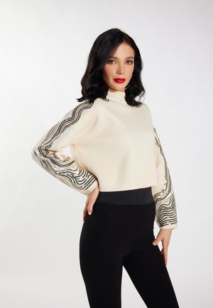 Donna con capelli neri che indossa un maglione color crema con motivi a onde di paillettes nere e oro sulle maniche e pantaloni neri a vita alta.