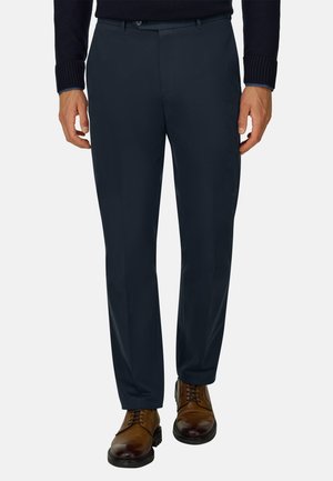 Homme portant un pantalon formel bleu marine foncé et des chaussures habillées en cuir marron, debout devant un fond uni.