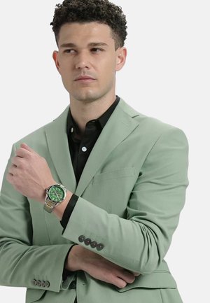 Roheline tekstuuriga bleiser, must särk, hõbedane käekell rohelise diali ja võrgurihmaga. Modell poseerib käed ristis, rõhutades kella detaile.