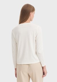 Long-sleeve cream cotton shirt with a round neckline, relaxed fit, and subtle seam detailing at the back. Worn with beige pants.