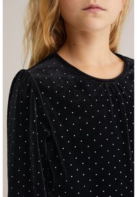 Melna samta blūze ar apaļu kakla izgriezumu un puff piedurknēm, dekorēta ar maziem baltajiem punktiem visā virsmā, lai pievienotu tekstūru.