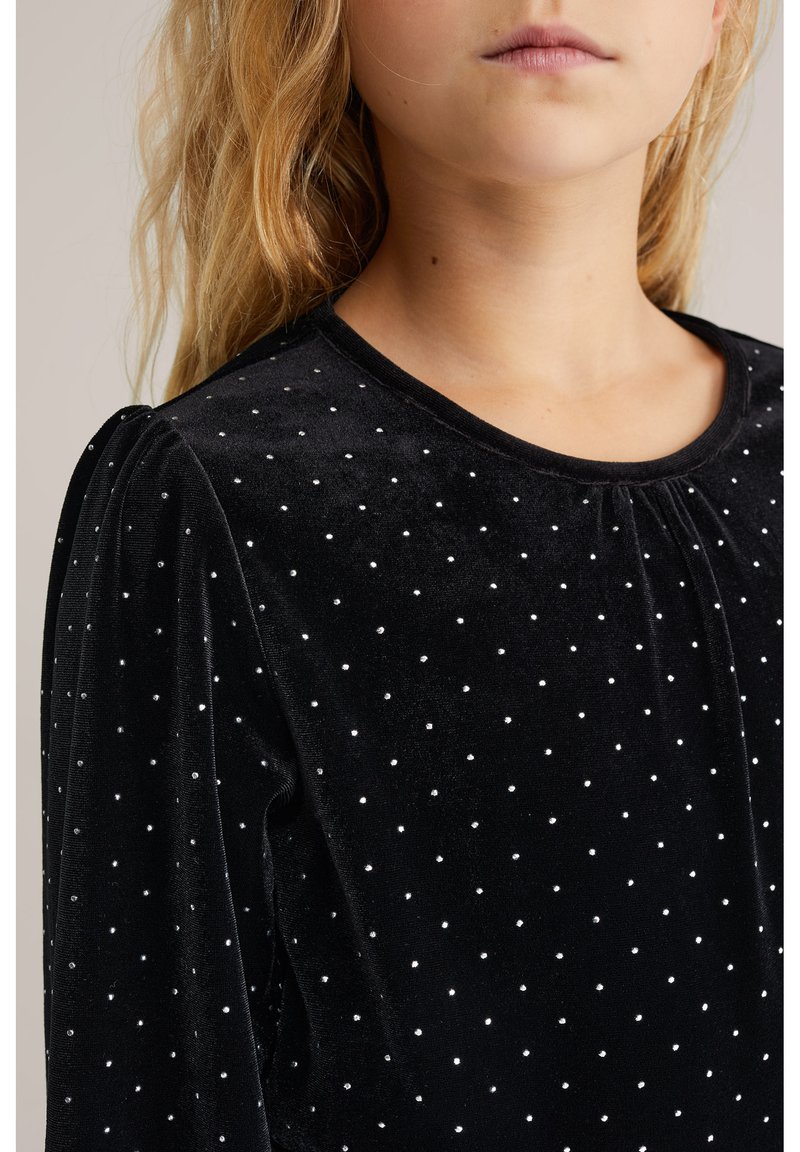 Črn sametast top z okroglo izrezom in nabranimi rokavi, okrašen z manjšimi belimi pikami po celotni površini za dodatno teksturo.