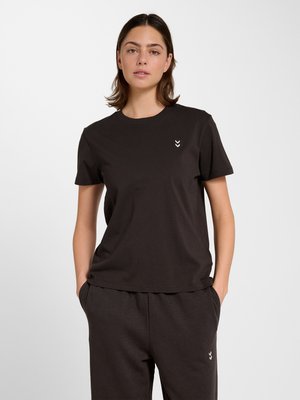 Kvinde iført sort kortærmet skjorte og matchende bukser, hænder i lommerne, stående mod en ensfarvet hvid baggrund.