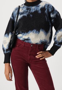 Strickpullover mit einem schwarzen, blauen und cremefarbenen Wolkenmuster; runder Ausschnitt. Burgunderfarbene Cordhosen mit strukturierten vertikalen Streifen und Vordertaschen.