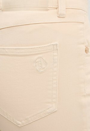 Primo piano della tasca posteriore di jeans in denim beige con logo ricamato discreto e rivetto di rame sulla cintura.