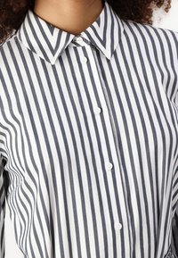 Pruhovaná košile s tenkými navy a bílými vertikálními pruhy, s špičatým límcem a zapínáním na knoflíky vpředu. Vyrobena z lehkého materiálu.