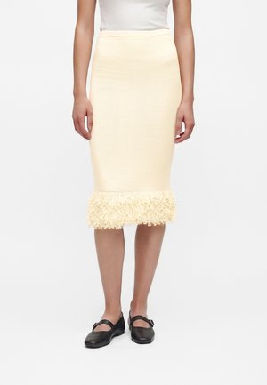 Femme portant une jupe crème mi-longue avec un ourlet à franges, un haut blanc et des ballerines noires style Mary Jane, debout devant un fond uni.