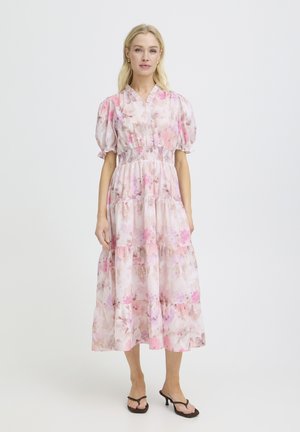 Frau trägt ein rosa-weißes Midikleid mit Blumenmuster, Puffärmeln und geraffter Taille, kombiniert mit schwarzen Sandaletten mit Absatz, vor einfachem Hintergrund.