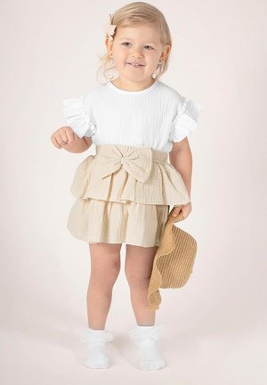 Biały, teksturowany top z krótkim rękawem i falbankami na rękawach, zestawiony z beżową, warstwową spódnicą z kokardą. Dziecko ma na nogach białe, falbankowe skarpetki i trzyma wiklinową torebkę.