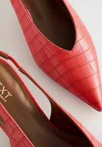 Tacchi slingback in ecopelle rossa effetto coccodrillo con punta affusolata e lati con intagli. Presentano un marchio dorato sulla soletta e un interno marrone liscio.