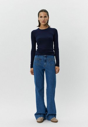 Jūras spēku tumši zila krekla ar garām piedurknēm apģērbs saskaņots ar augsto jostu gaiši ziliem platajiem džinsiem. Džinsiem ir aizmugures kabatas un brīvs piegriezums, ko papildina bēši apavi.