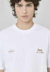 T-shirt en coton blanc avec le texte brodé marron "gagnant" et "Meilleur de sa catégorie". Col rond classique et manches courtes. Texture lisse.