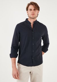 Camisa de manga larga azul marino oscuro con cuello mao, cierre de botones en el frente y detalles de manga enrollada. La tela tiene una textura suave.