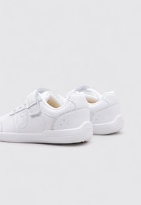 Zapatillas blancas hechas de material sintético con una textura suave. Presentan una correa de velcro, un detalle de logo sutil y un diseño minimalista.