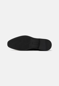 Suela de zapato negra con goma texturizada, que presenta un patrón de líneas diagonales y un leve tacón. Punta redondeada y bordes suaves.