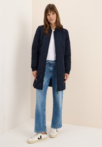 Giacca lunga imbottita blu navy con polsini a coste e zip frontale, abbinata a jeans a gamba larga azzurri e scarpe bianche con dettagli neri.
