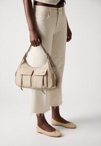 Μπεζ σουέτ τσάντα hobo με διπλές μπροστινές τσέπες, χρυσά μεταλλικά στοιχεία και μία μόνο λουρί ώμου, κρατημένη στο χέρι κοντά σε ανοιχτόχρωμα cropped παντελόνια.