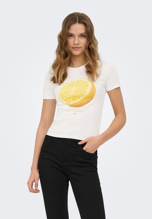 Joven mujer con cabello ondulado que lleva una camiseta blanca con un gran gráfico de limón amarillo, combinada con pantalones negros, de pie frente a un fondo liso.