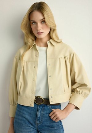 Blondynka w kremowej zapinanej na guziki kurtce, białej koszuli, niebieskich dżinsach z brązowym paskiem, stojąca na tle białej ściany.