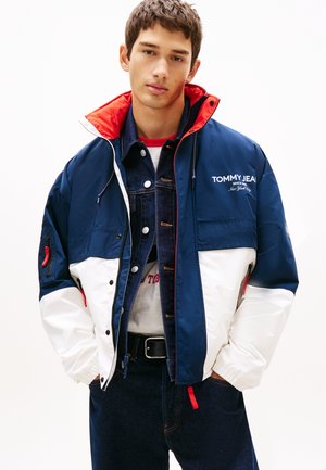 Fiatal férfi, aki egy tengerészkék, fehér és piros Tommy Jeans dzsekit visel egy farmerdzseki és farmernadrág felett, kezét zsebre dugva, előre nézve.