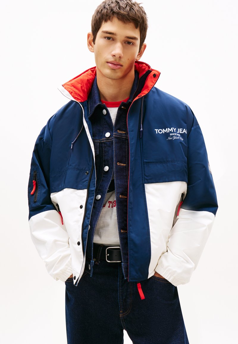 Νέος άνδρας που φοράει ένα μπουφάν Tommy Jeans σε μπλε ναυτικό, λευκό και κόκκινο χρώμα πάνω από ένα τζιν μπουφάν και τζιν, με τα χέρια στις τσέπες, κοιτάζοντας μπροστά.