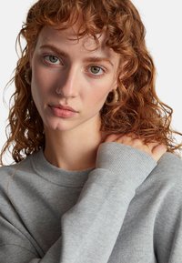 Felpa grigia con scollo e polsini a coste. Tessuto strutturato, vestibilità morbida. Il modello si tocca la spalla, mostrando orecchini e trucco naturale.