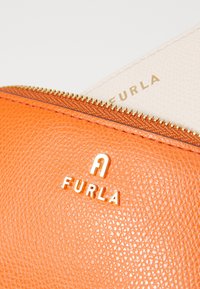 Narančasta kožna torbica s zlatnim zatvaračem, koja se odlikuje teksturiranom površinom i utisnutim zlatnim logotipom "FURLA". Unutarnji materijal je mekan bež.