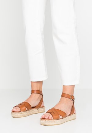 Persona che indossa pantaloni bianchi corti e sandali platform in pelle marrone con cinturino alla caviglia e suola espadrillas intrecciata su un pavimento bianco.