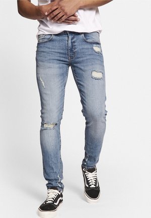 Jean skinny en denim bleu clair avec des détails usés, comprenant des déchirures et des bords effilochés. Associés à des baskets noires et blanches.