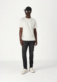 Bijela majica kratkih rukava, tamne traperice, bijele tenisice i crna kapa. Sunčane naočale s laganim okvirima. Jednostavan, ležeran outfit.