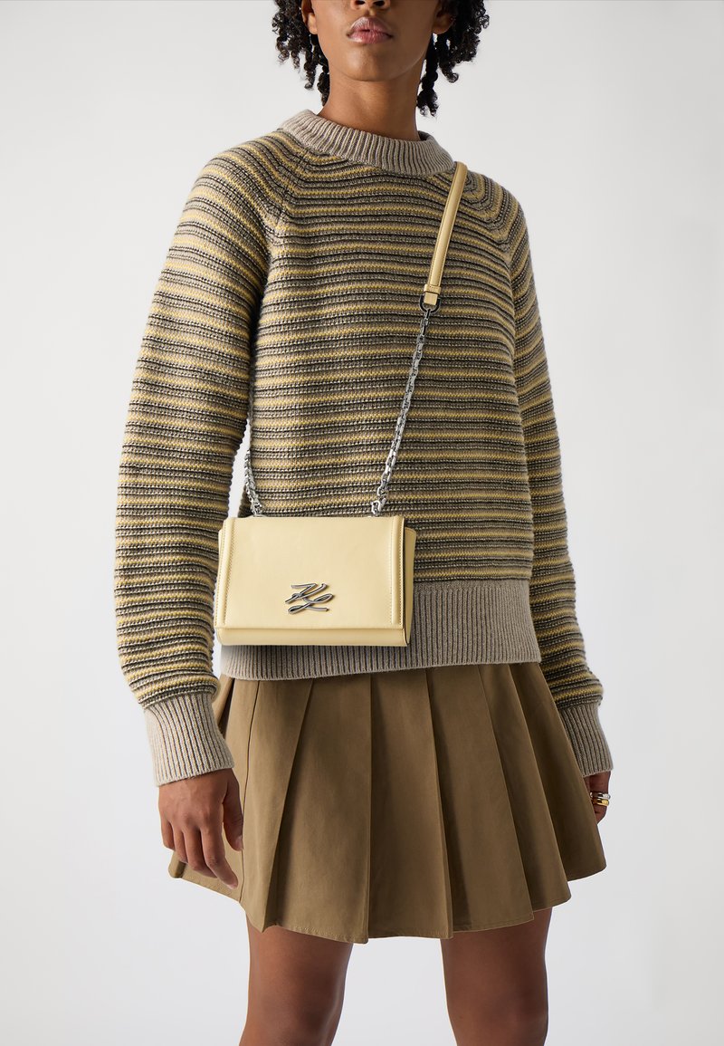 Gul crossbody-veske med kjedehank, glatt skinnfinish og en metallisk logo. Båret med en stripete strikkegenser og plisserte skjørt.