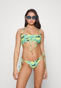 Conjunto de biquíni verde com padrão de frutas. O top tem laços ajustáveis e um centro franzido, enquanto a parte de baixo apresenta laços laterais.