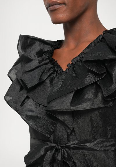 Mustarik bluus, millel on volangiga kaelus, tekstureeritud satiinkangas, elastsed äärised ja sidekoht. Rõhk on kihilistelt volangidelt ja sädelevalt pinnalt.