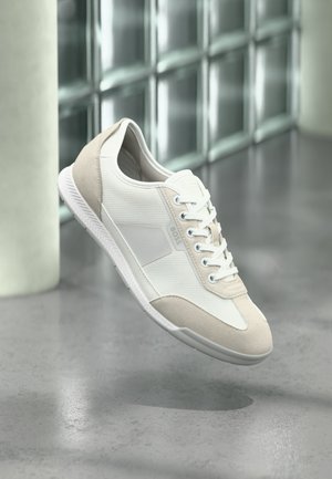 Zapatilla casual blanca y beige con cordones, paneles texturizados y el logo "BOSS" en el lateral, flotando sobre un suelo de hormigón pulido.