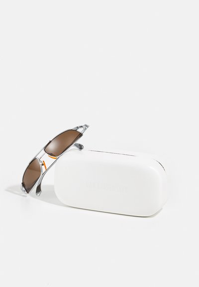 Des lunettes de soleil avec des verres teintés marron et des montures en métal argenté, placées à côté d'un étui rectangulaire blanc avec un texte en relief.