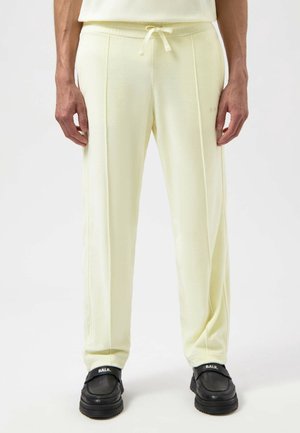Homme portant un pantalon crème avec un cordon de serrage et des coutures à l'avant, ainsi que des chaussures noires slip-on avec le logo blanc "BALR." sur chaque chaussure.