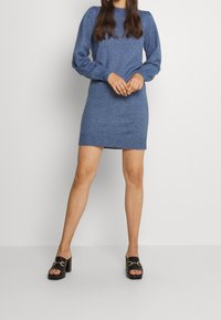 Abito blu a maglia, a maniche lunghe, con una forma aderente e orlo a coste. Sandali neri con tacco a blocco e dettagli in oro.