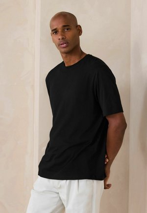 Mies, jolla on musta lyhythihainen paita ja valkoiset housut, seisoo kädet selän takana beigenväristä teksturoitua seinää vasten.