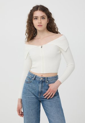 Biały prążkowany crop top z dekoltem off-the-shoulder i zamkiem błyskawicznym z przodu, w połączeniu z niebieskimi dżinsami o wysokim stanie.