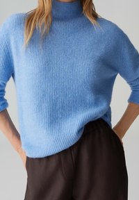 Personne portant un pull en maille bleu clair doux avec un col montant et un pantalon ample marron foncé, les mains dans les poches.