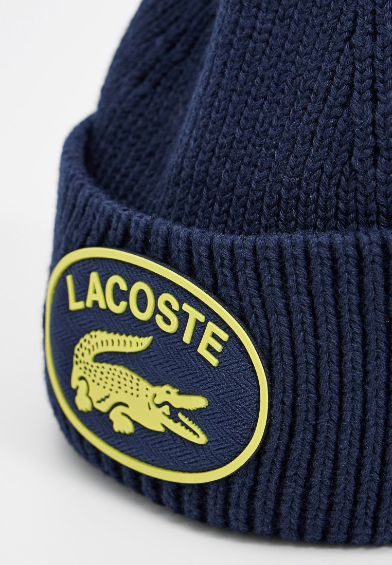Marineblauer Strickbeanie mit einem strukturierten Rippendesign, versehen mit einem gelben ovalen Abzeichen, auf dem "LACOSTE" und ein Krokodil-Logo zu sehen sind.