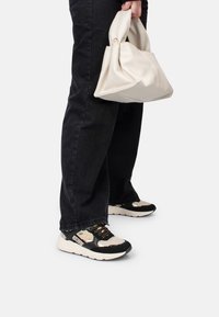 Jeans noirs, sac à main crème et baskets noir et blanc avec des panneaux en mesh et lisses, des accents dorés et des détails texturés sur le dessus.